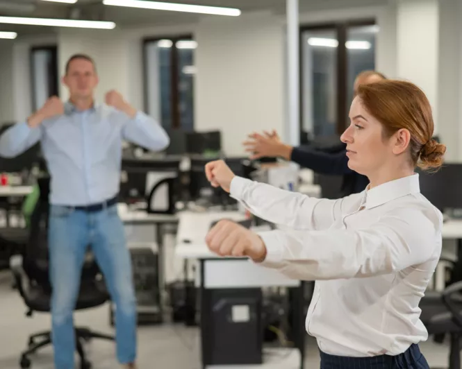Büroangestellte führen gemeinsam leichte Dehn- und Fitnessübungen in einem modernen Büro durch. Im Vordergrund eine Frau in weißer Bluse, die konzentriert ihre Arme streckt, während im Hintergrund weitere Kollegen aktiv teilnehmen.
