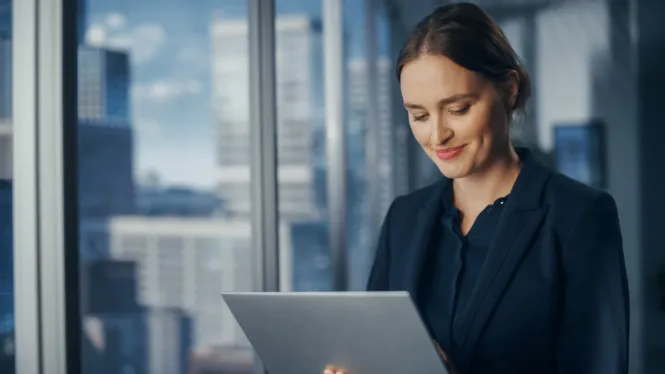 Frau mit schwarzen Haaren in Business Outfit schaut lächelnd auf ein Tablet.