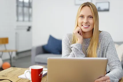 Lächelnde blonde Frau arbeitet am Laptop, Kaffeetasse & Notizbuch auf dem Tisch.