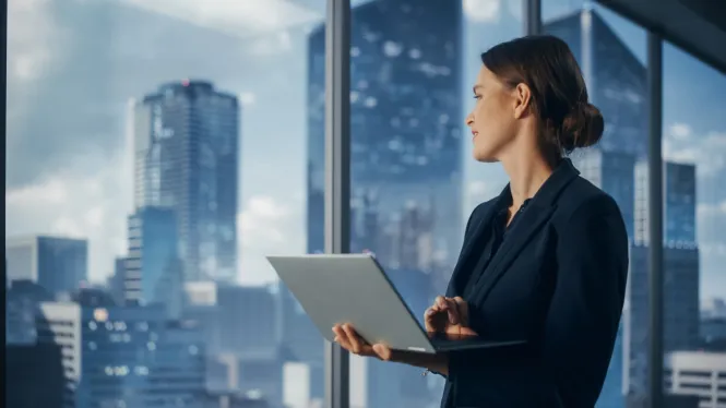 Eine Frau im Business Outfit mit Laptop in der Hand schaut aus dem Fenster.